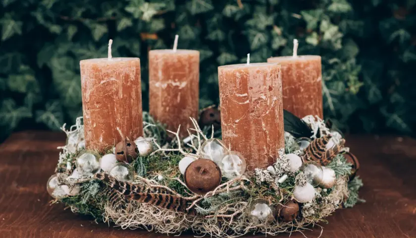 A rustic Advent wreath with four brown candles, moss, feathers, and glass ornaments on a wooden table.