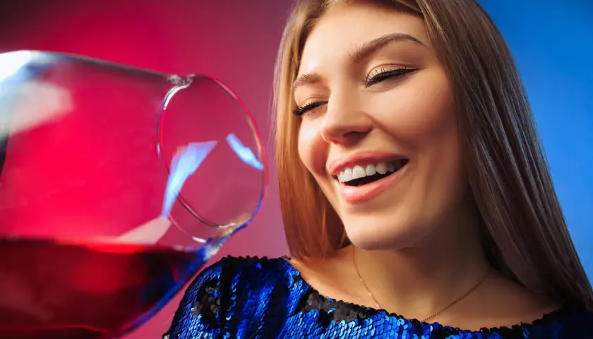 Uśmiechnięta kobieta w błyszczącej sukience, z kieliszkiem wina. Czy po botoksie można pić alkohol?