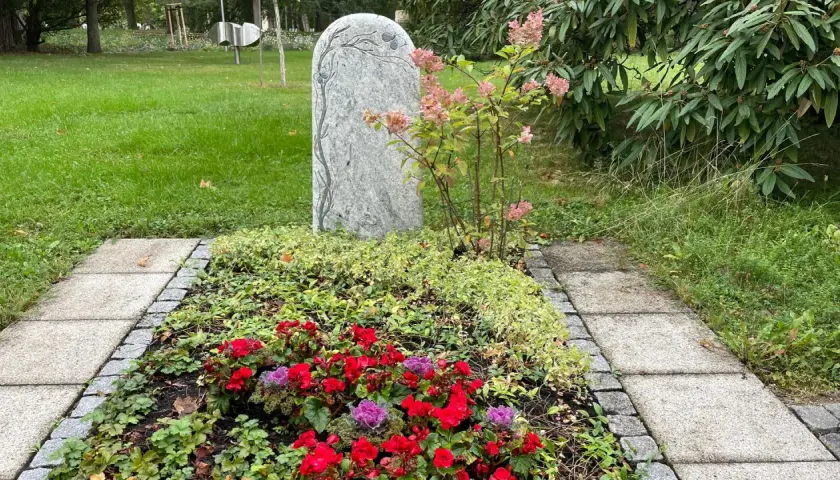 Ein Grabstein mit Baumgravur, umgeben von bunten Blumen und immergrünen Pflanzen. Dauerhafte Grabbepflanzung Beispiele für eine liebevolle Gestaltung.