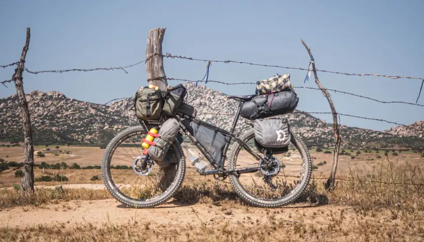 Bicicleta lista para bike packing, cargada con alforjas y bolsas, apoyada en un poste de alambre de espino, con paisaje montañoso al fondo.