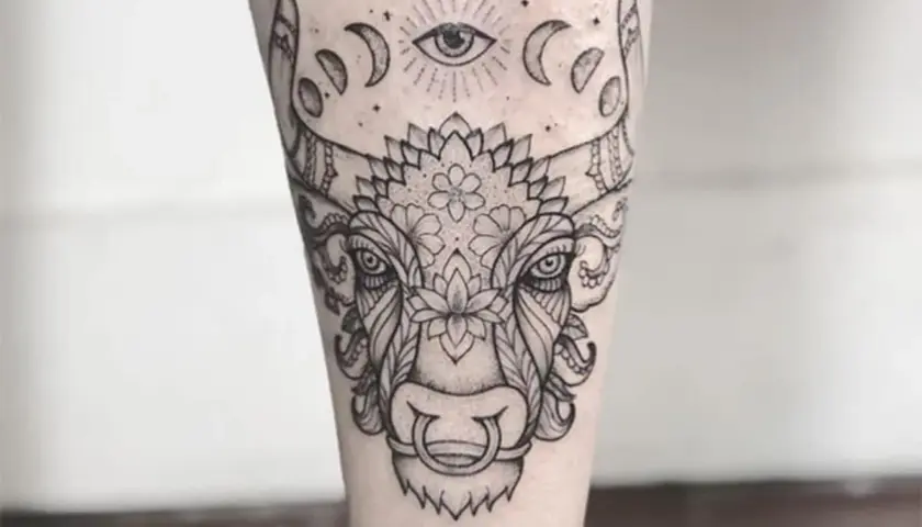 Un detallado tatuaje taurino con un ojo místico y fases lunares sobre la cabeza de un toro adornado con motivos florales y ornamentales.