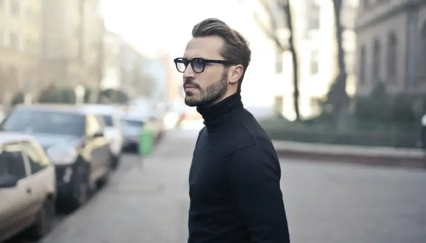Elegancki mężczyzna w okularach i czarnym golfie, zamyślony, jakby planował zagrywki zakochanego faceta.