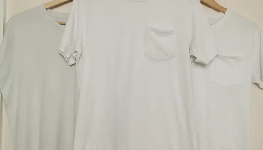 Trois t-shirts blancs sur cintres en bois. Le t-shirt du milieu a une poche poitrine. Idéal pour apprendre comment blanchir un pull blanc.