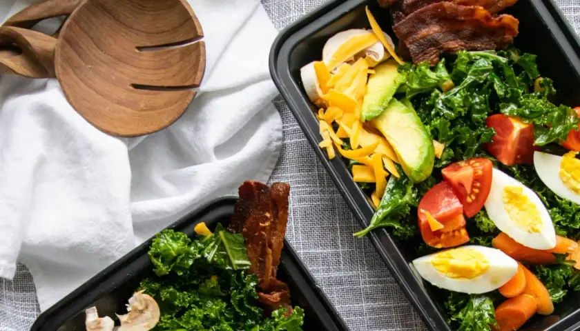 Zwei Behälter mit leckeren low carb meal prep Salaten, gefüllt mit Grünkohl, Avocado, Tomaten, Eiern, Käse und Speck.