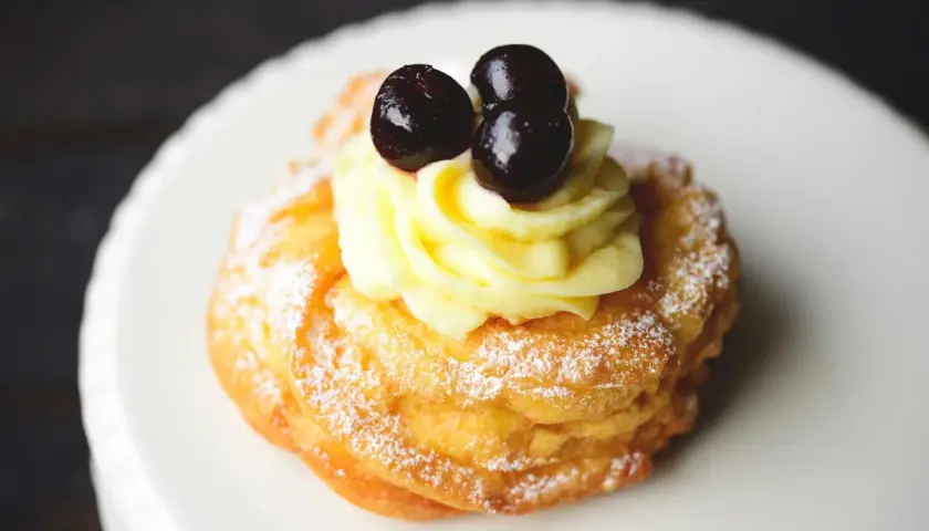 Zeppole fritte gonfie, soffice pasta con crema pasticcera e ciliegie candite, spolverate di zucchero a velo.