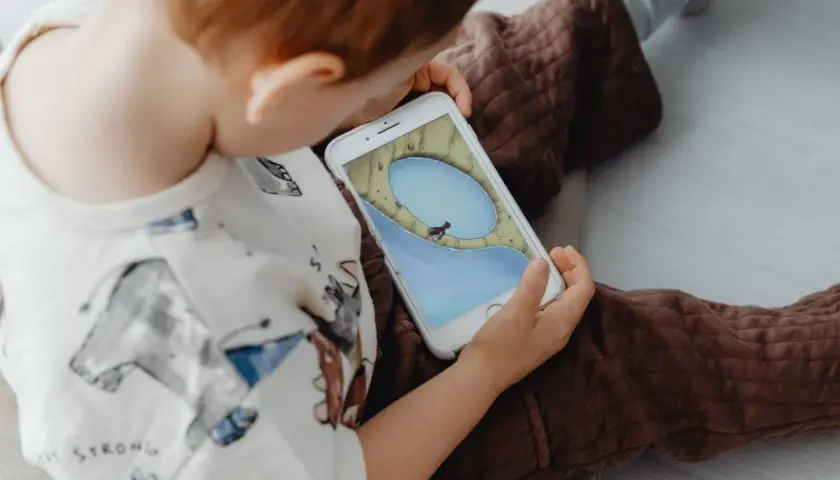Un niño, absorto en su móvil, parece un niño adicto al móvil. La pantalla muestra un juego con un personaje en un paisaje acuático.