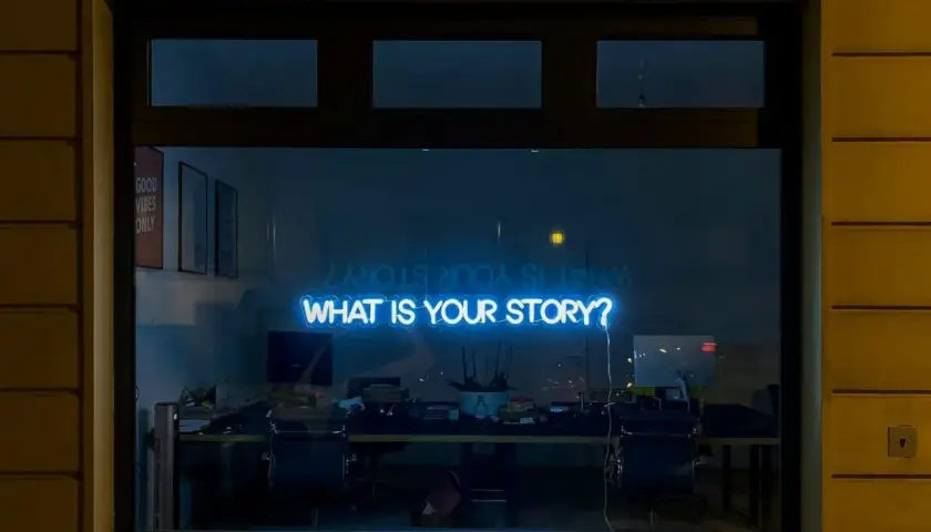 Neonskylt med texten "WHAT IS YOUR STORY?" lyser blått i ett fönster, som en inbjudan till en kurs i skrivande.