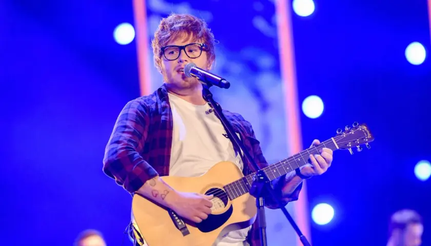 Ed Sheeran w okularach gra na gitarze akustycznej i śpiewa do mikrofonu na niebieskiej scenie.