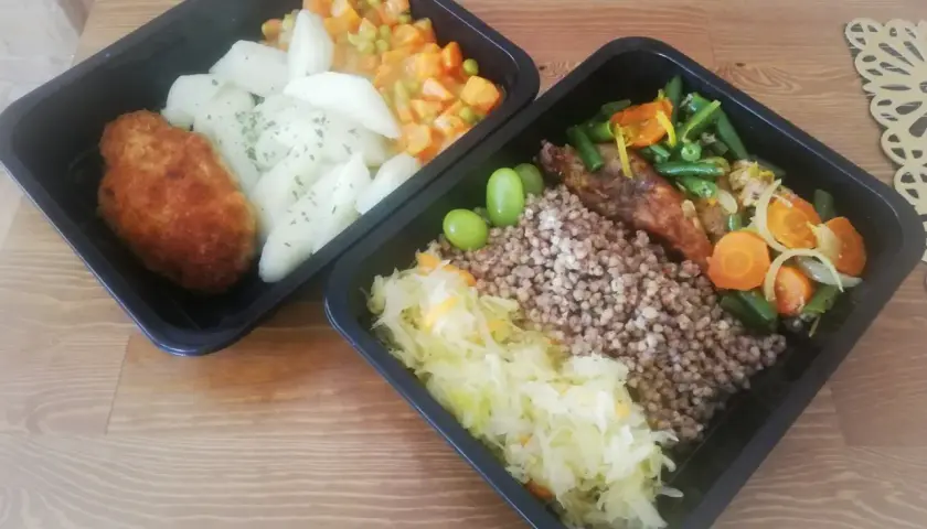 Dwa czarne pojemniki z jedzeniem: kotlet, ziemniaki, warzywa, kasza gryczana, surówka i winogrona.