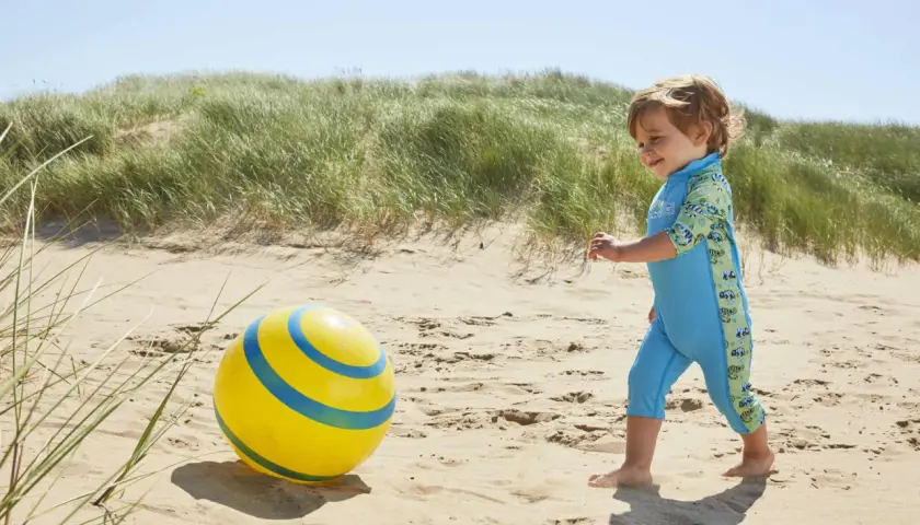 Chłopiec w błękitnym stroju kąpielowym dla niemowląt bawi się na plaży z żółtą piłką.