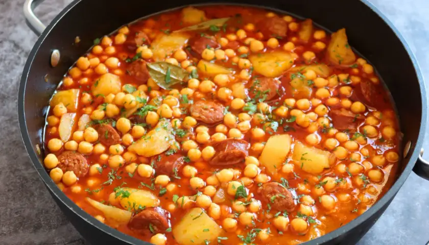 Un plato humeante de garbanzos con chorizo arguiñano, patatas y perejil picado en una sartén negra.