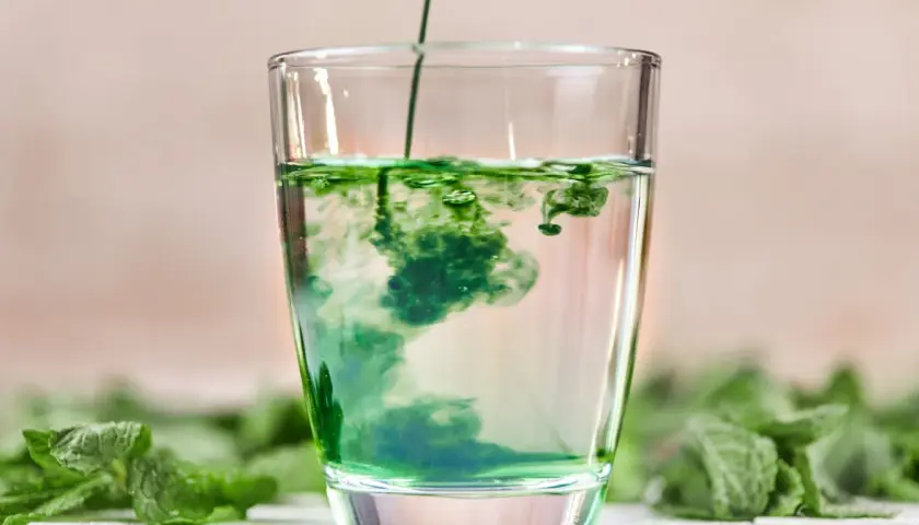 Zielony płyn rozpuszcza się w szklance wody, tworząc wiry. To naturalny chlorofil do picia, otoczony świeżymi liśćmi mięty.