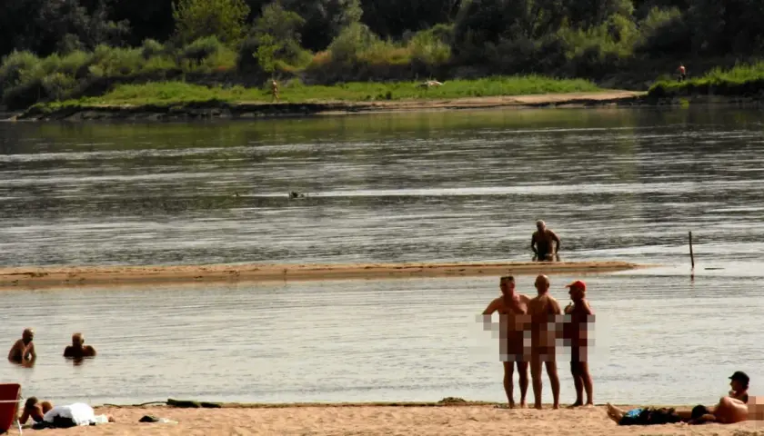 Ludzie opalają się i kąpią na plaży nudystów. Woda jest spokojna, a na brzegu widać kilka osób.