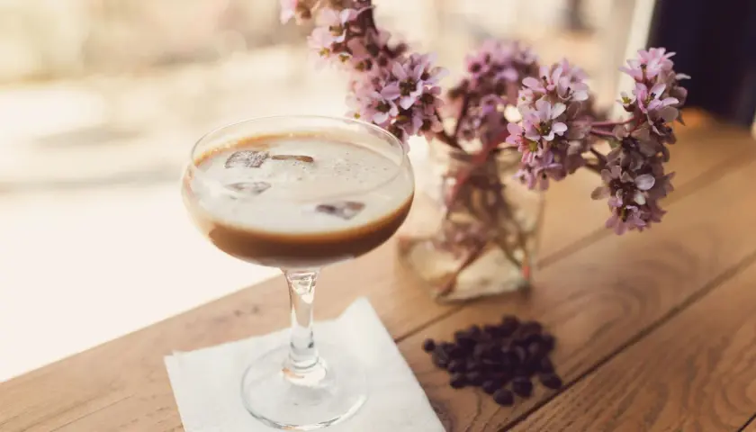 Ein cremiger Eiskaffee-Cocktail mit Eiswürfeln, daneben zarte rosa Blüten und Kaffeebohnen auf Holztisch.