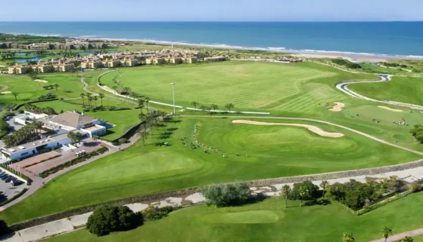 Vista aérea del campo de golf del hotel Barceló Costa Ballena, con el mar azul al fondo y edificios residenciales.