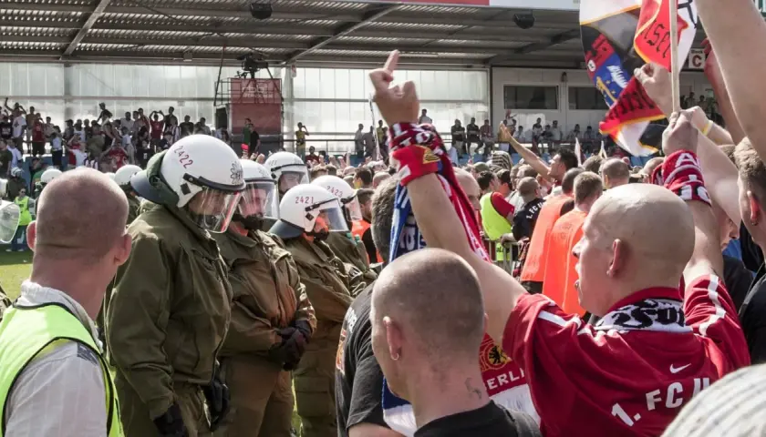 Policja w pełnym rynsztunku stoi naprzeciwko grupy kibiców, wśród których widać ultrasi z flagami i szalikami.