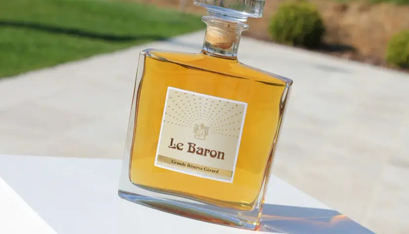 Bouteille de "Le Baron Grande Réserve Gérard", un spiritueux ambré, posée sur un socle blanc, avec un arrière-plan flou évoquant un jardin et un chemin, rappelant l'élégance d'un chateau vaudois.