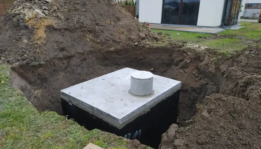 Zbiornik na deszczówkę czeka na podłączenie do rynny. Wykop jest gotowy, a betonowa pokrywa z kominkiem widoczna.