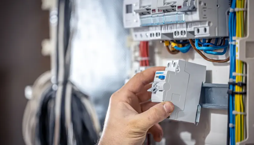 Dłoń instaluje bezpiecznik w rozdzielni elektrycznej, widoczne przewody i inne bezpieczniki.