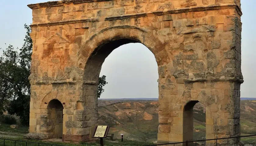Imponente arco romano de piedra, con dos arcos menores a los lados, se alza sobre un paisaje de colinas verdes.