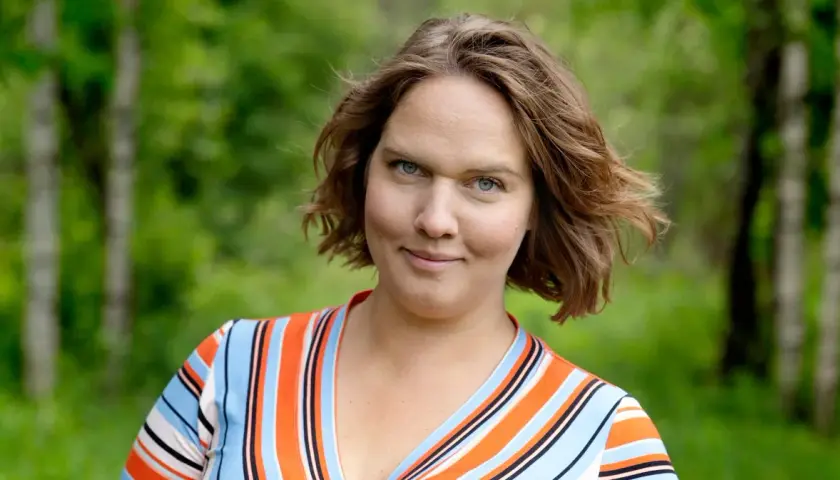 Underbara Claras leende möter kameran i en randig klänning, med en lummig skog i bakgrunden.