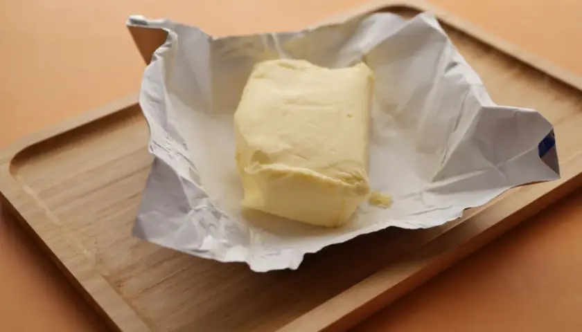 Kostka masła w folii na drewnianej desce. Czy masło można mrozić? Tak, można je zamrozić, by zachować świeżość.