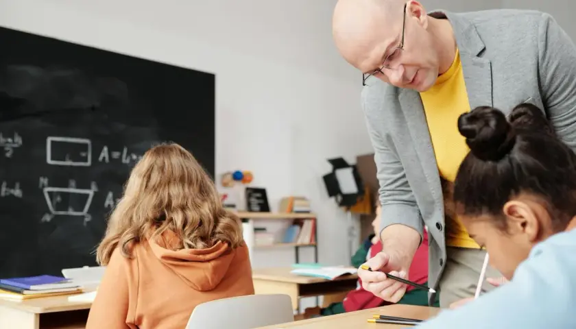 Nauczyciel pomaga uczniom przy biurkach w klasie, na tablicy widać wzory matematyczne.