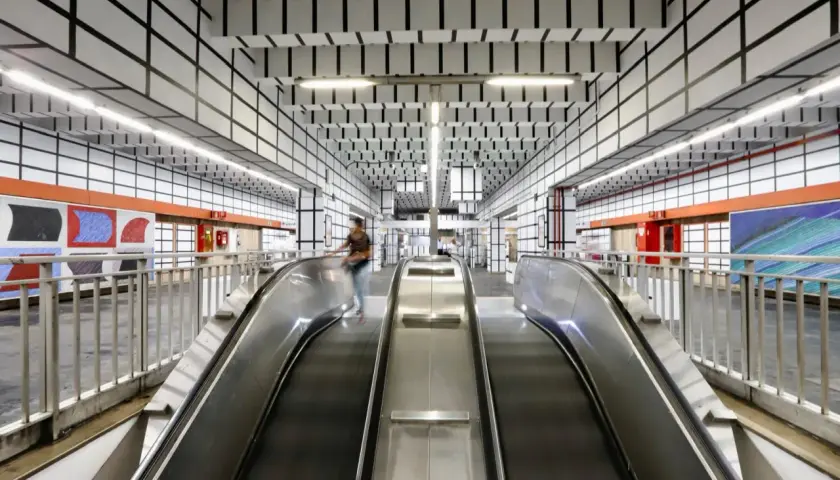 Scale mobili nella moderna stazione metro Vittorio Emanuele a Roma, con soffitto a griglia nera su bianco e opere d'arte colorate alle pareti.