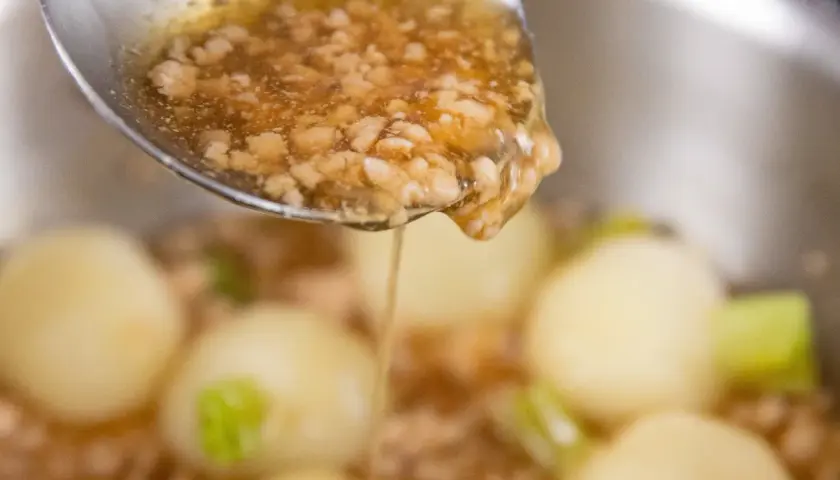 A spoon pours a thick, savory ankake sauce over tender dumplings and vegetables in a pot.