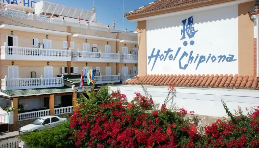 Fachada del Hotel Chipiona con balcones blancos, flores rojas y un coche aparcado.