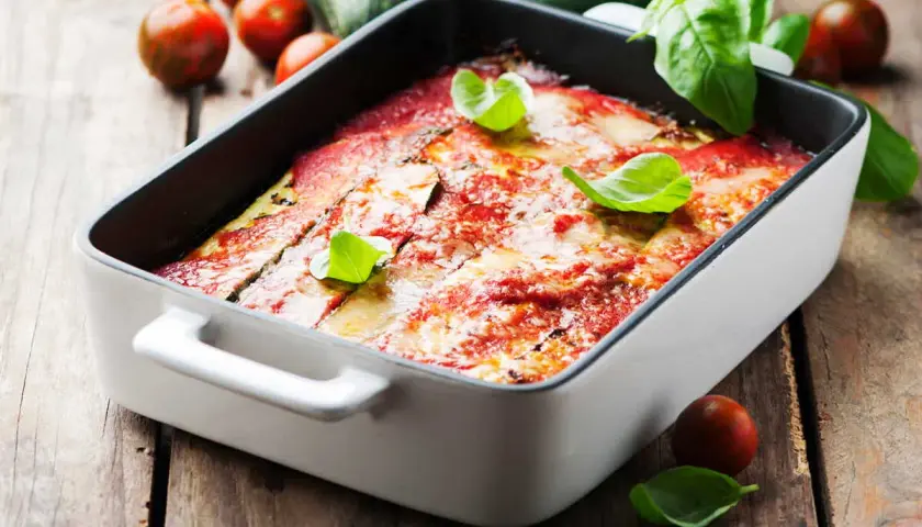 Lasagne courgette gratinée, garnie de basilic frais, sur fond de bois rustique avec des tomates cerises et des courgettes.