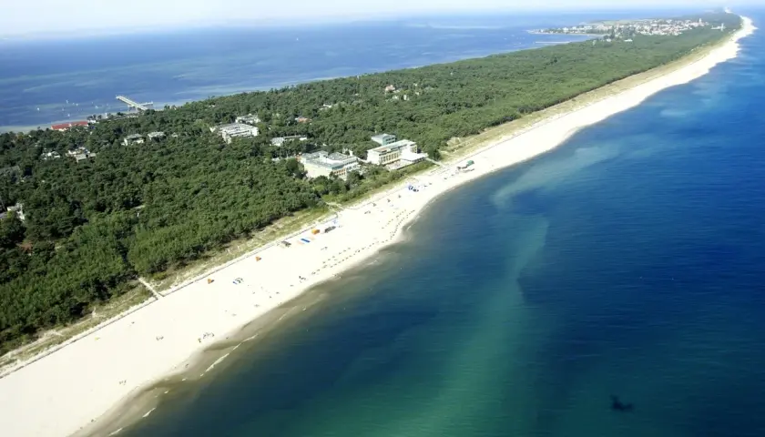 Jurata plaża z lotu ptaka: szeroka, piaszczysta plaża, sosnowy las i budynki hotelowe nad turkusowym morzem.