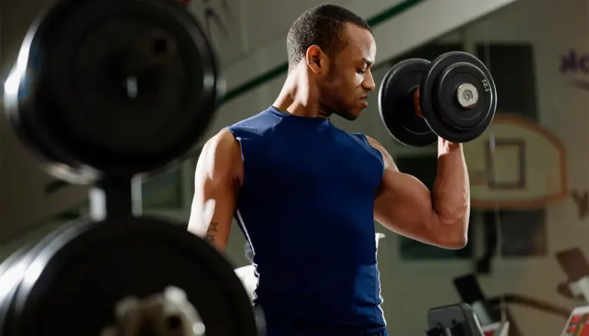 Mężczyzna ćwiczy biceps z hantlem. W tle widać sprzęt do ćwiczeń.