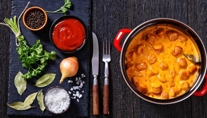 Pyszne parówki w sosie śmietanowym – idealny pomysł, co zrobić z parówek na obiad. Obok składniki: cebula, ketchup, przyprawy.