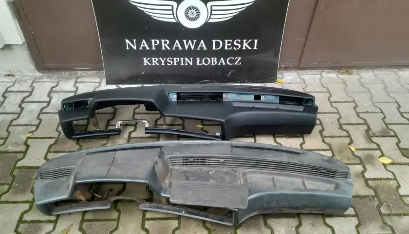 Dwa kokpity samochodowe, jeden czarny, drugi szary, leżą na chodniku przed tablicą z napisem "NAPRAWA DESKI".