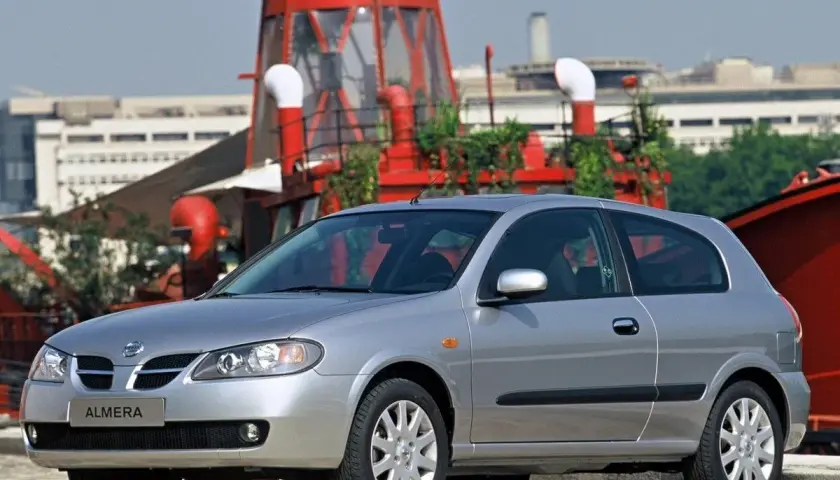 Srebrny Nissan Almera stoi przed czerwoną, industrialną konstrukcją.
