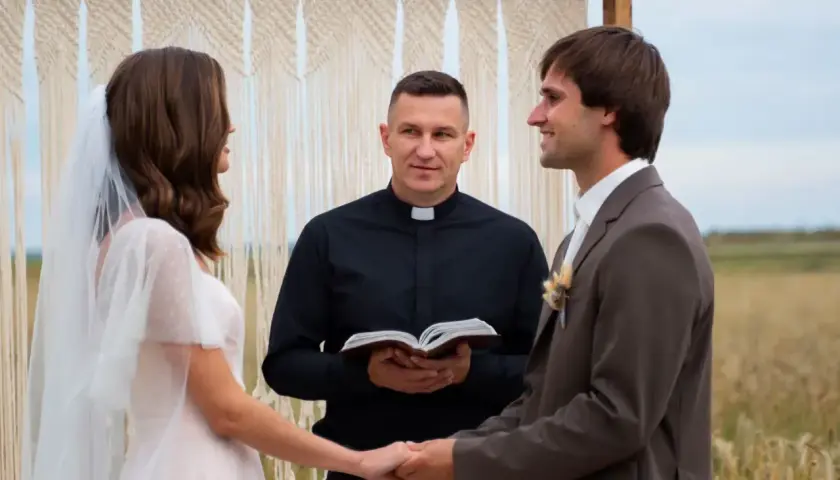 Ksiądz na ślubie, trzymając Biblię, błogosławi parę młodą. Panna młoda w białej sukni i welonie, pan młody w garniturze.