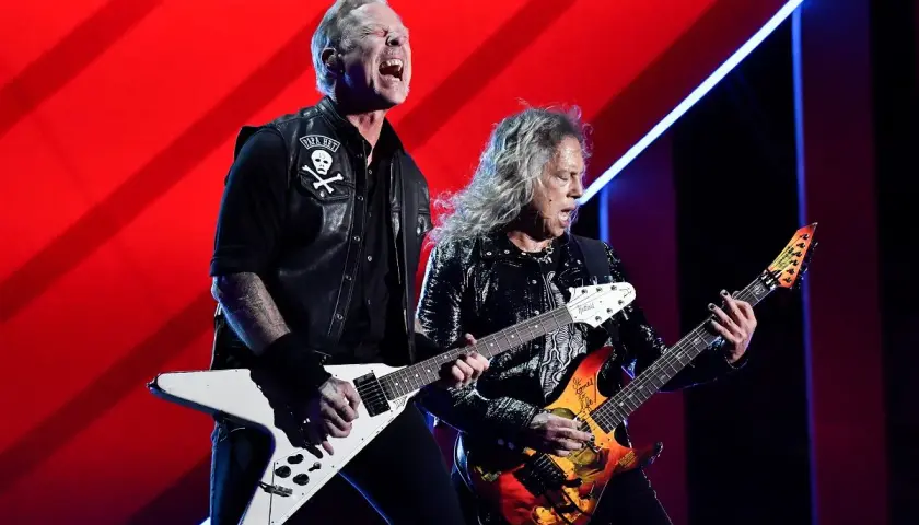 Dwóch gitarzystów zespołu Metallica gra na scenie na tle czerwonej kurtyny.