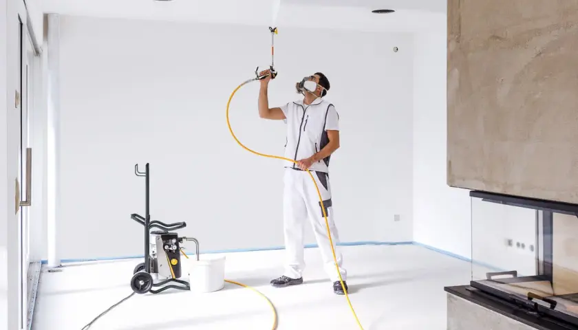 Pittore con maschera protettiva usa una pistola per vernice a spruzzo per dipingere il soffitto di una stanza vuota.