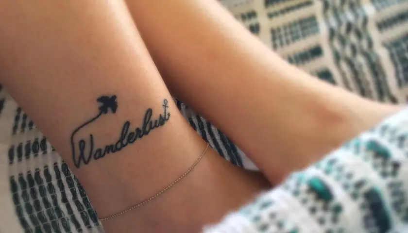 Delikatny tatuaż podróżniczy na nadgarstku: napis "Wanderlust" z samolotem i kotwicą.