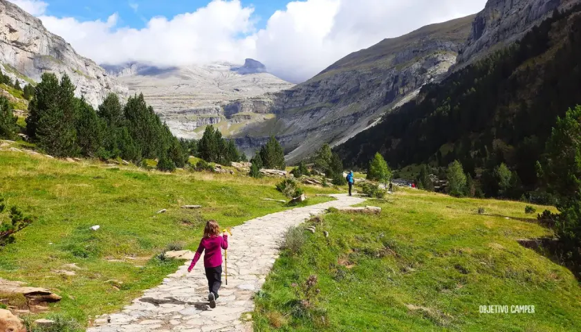 Niños disfrutan de la ruta cola de caballo, un sendero de piedra serpentea por un valle montañoso con pinos y cielos nublados.