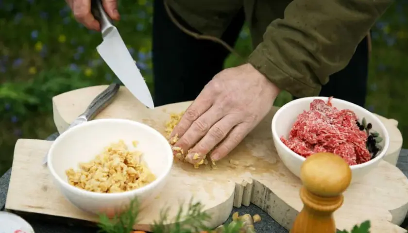 Förbereder eko mat utomhus. En hand hackar ingredienser på en träskärbräda bredvid skålar med köttfärs och en annan ingrediens.