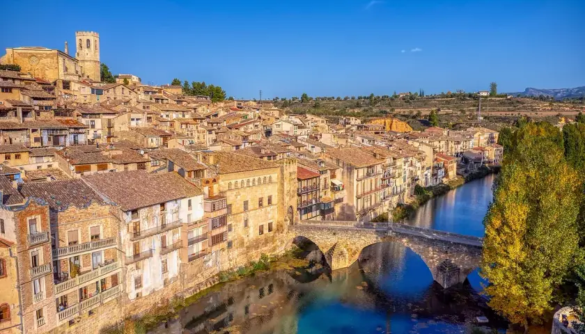 Pueblo con casas de tejas de barro y un puente de piedra sobre un río azul, evocando la toscana española.