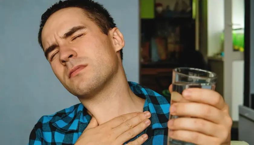 Mężczyzna z zamkniętymi oczami trzyma szklankę wody i dotyka gardła.