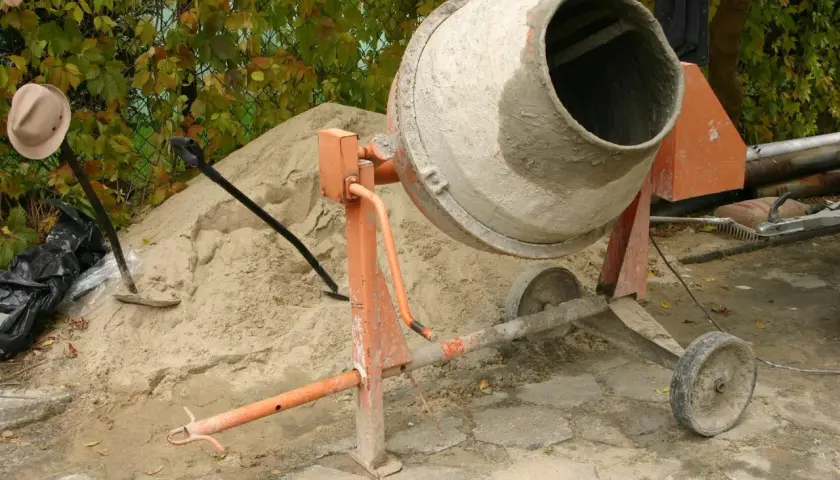 Betoniarka 120l czeka na cement. Obok piasek, łopata i kapelusz.
