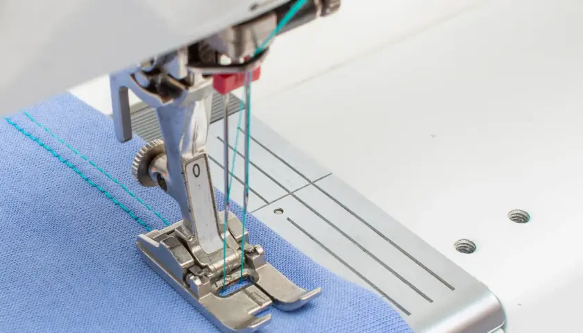 Close-up of a sewing machine needle plate and foot, demonstrating how to use a double needle on blue fabric with turquoise thread.
