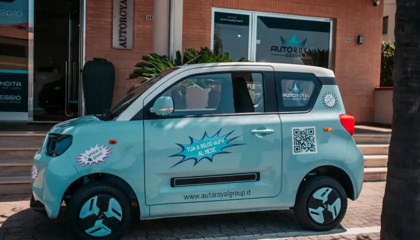 Macchina piccola 4 posti azzurra, con adesivi promozionali "TUA A 99,00 euro AL MESE" e QR code.