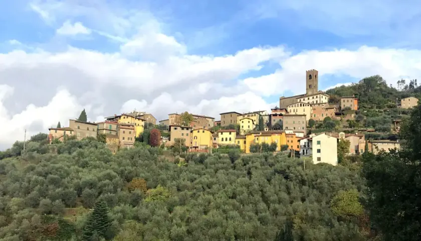 Un pittoresco borgo a Buggiano, con case colorate arroccate su una collina ricoperta di ulivi, sotto un cielo azzurro con nuvole bianche.