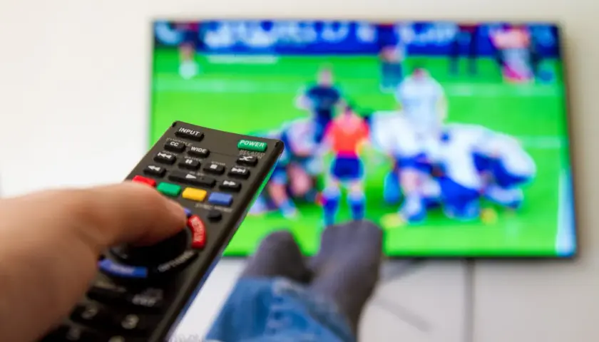 Dłoń trzymająca pilot do telewizora, na ekranie mecz piłkarski.
