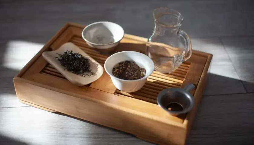 Zestaw do gongfu cha: drewniana taca, liście herbaty, czajniczek, miseczki i sitko.